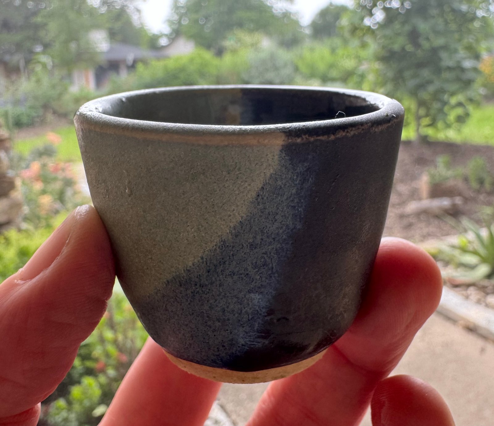 O-Choko β Handcrafted Ceramic Sake Cup A handcrafted ceramic o-choko sake cup with a green and dark glaze, resting on a blue glass surface outdoors